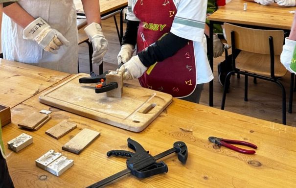 Eine Person schneidet Holz auf einem Tisch, umgeben von Werkzeugen und Materialien.