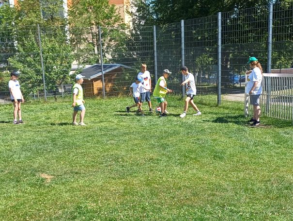 Sechs Kinder spielen auf einer Wiese Fußball, umgeben von einem Zaun und Bäumen.
