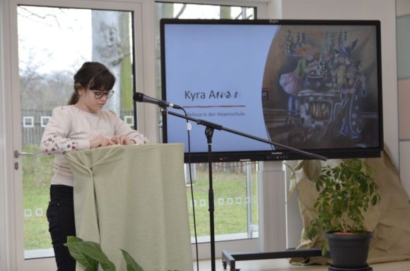 Ein Mädchen steht hinter einem Podium, während eine Präsentation im Hintergrund läuft.