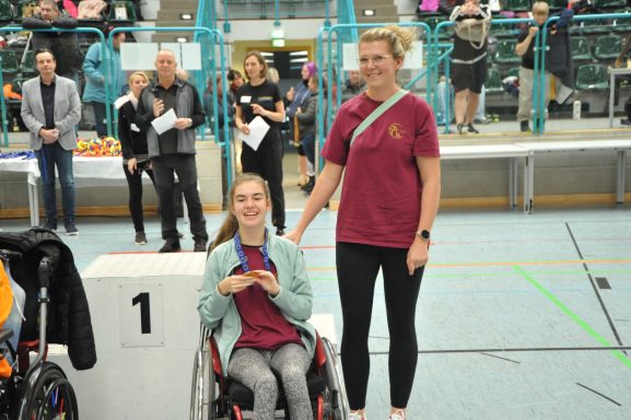 Zwei Personen stehen in einem Sportumfeld, eine im Rollstuhl mit Medaille, lächelnd.
