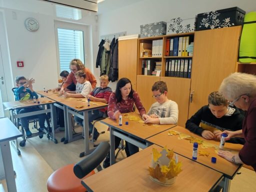 Kinder und Erwachsene sitzen an Tischen und basteln in einem Klassenraum.