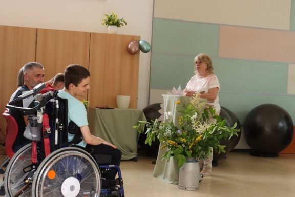 Eine Gruppe Menschen in einem Raum mit Pflanzen und Fitnessbällen, einer sitzt im Rollstuhl.