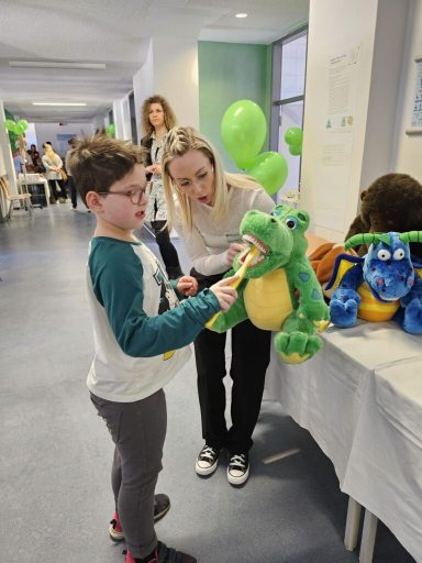 Ein Kind interagiert mit einem Plüschtier, während eine Frau ihm hilft. Grüne Ballons im Hintergrund.