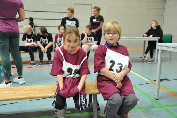 Zwei Kinder in lila Trikots mit Nummern sitzen auf einer Holzbank in der Turnhalle.