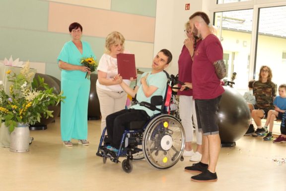 Eine Person im Rollstuhl erhält eine Auszeichnung, umgeben von Personen und Dekorationen.