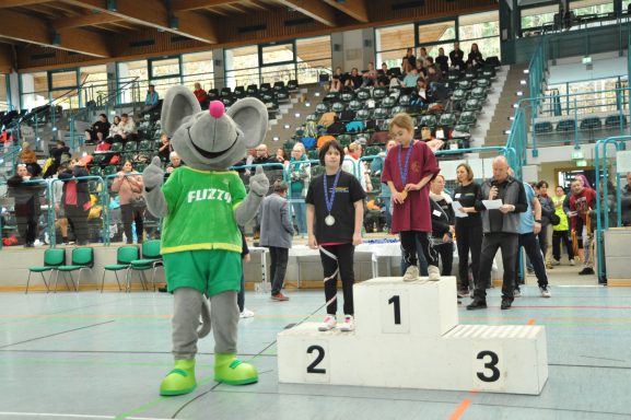 Ein Maskottchen steht neben Siegern auf einem Podest in einer Sporthalle.