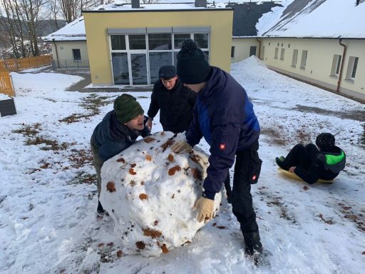 Fünf Personen bauen einen großen Schneeball auf einem schneebedeckten Gelände.