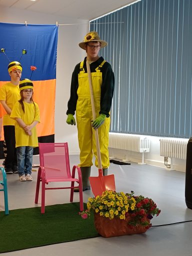 Person in gelber Kleidung mit Schaufel und Blumen, umgeben von kleinen Stühlen und Kindern.