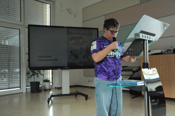 Junger Mann mit Brille steht an einem Rednerpult vor einem Bildschirm.