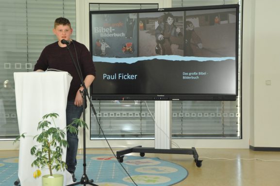 Ein junger Mann spricht an einem Tisch vor einem Bildschirm mit Präsentation.
