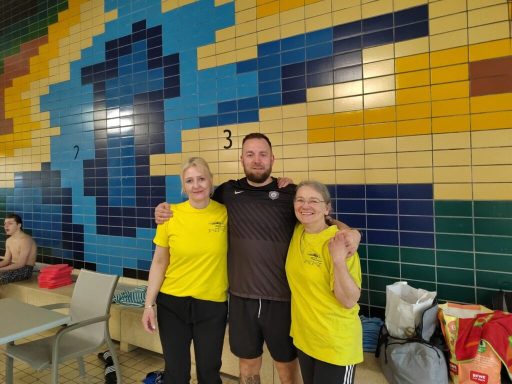 Drei Personen in gelben T-Shirts stehen neben einer bunten Wand im Schwimmbereich.
