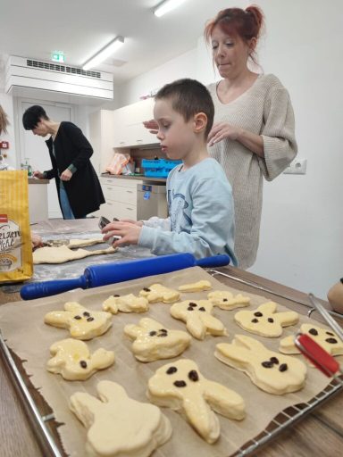 Ein Junge rollt Keksteig aus, während Erwachsene im Hintergrund helfen. Plätzchen in Form von Tieren.