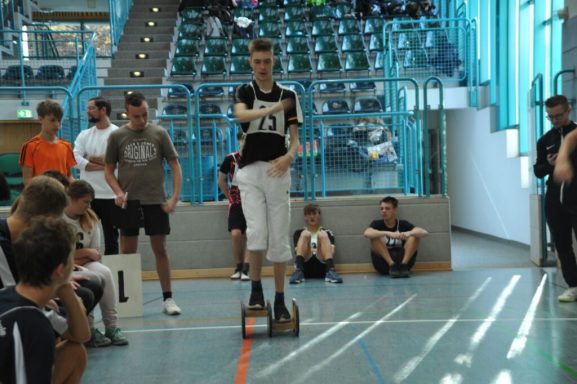 Jugendlicher auf einem Skateboard in einer Sporthalle, umgeben von Zuschauern.