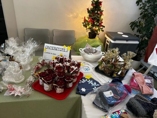 Tisch mit verschiedenen handgefertigten Geschenken und Dekorationen, Weihnachtsbaum im Hintergrund.