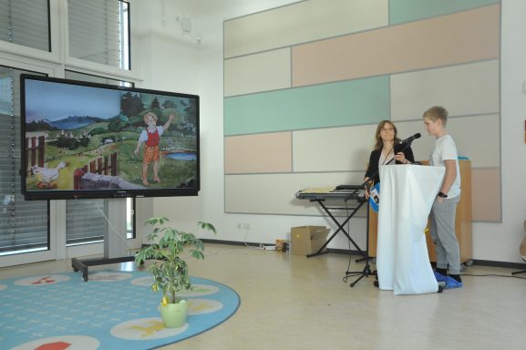 Ein Junge und ein Mädchen stehen an einem Rednerpult, während ein Bildschirm hinter ihnen leuchtet.