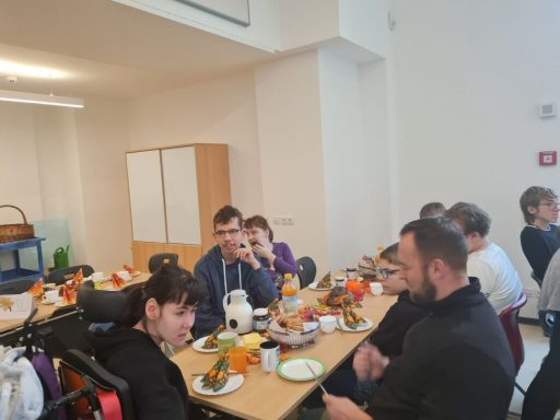 Gruppe von Menschen beim Essen in einem Raum mit Tischen und Stühlen.
