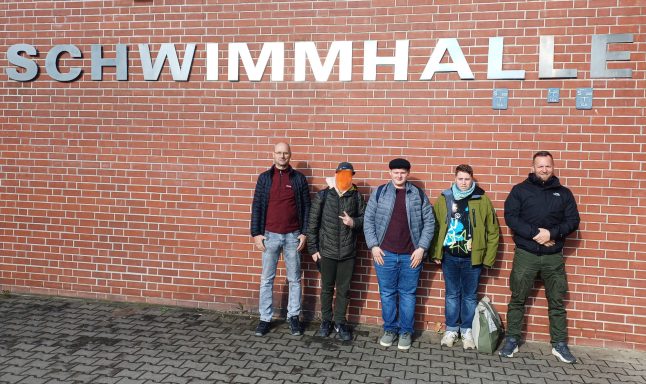 Fünf Personen stehen vor einer Schwimmhalle mit grauer Wand und Schriftzug.