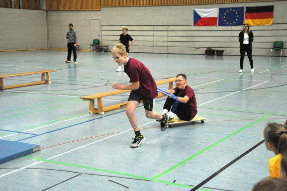 Zwei Kinder in Sportbekleidung, eines läuft, das andere sitzt auf einem Rollbrett.