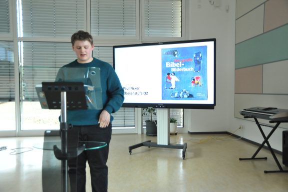 Junger Mann steht an einem Rednerpult vor einem Bildschirm mit Präsentation.