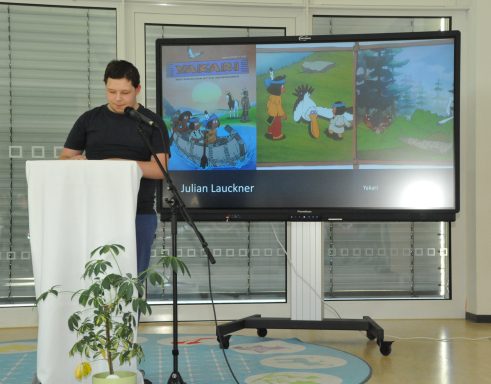 Junger Mann liest vor Publikum an einem Tisch mit einem Bildschirm im Hintergrund.