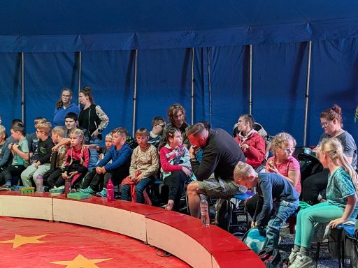 Kinder sitzen gespannt in einem Zirkus, umgeben von bunten Dekorationen.
