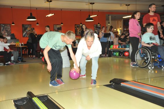 Zwei Personen beugen sich über eine Bowlingbahn, während andere im Hintergrund bowlen.