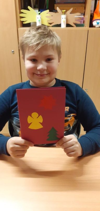 Ein Junge hält eine bastelnde Weihnachtskarte mit einem Engel und einem Baum.