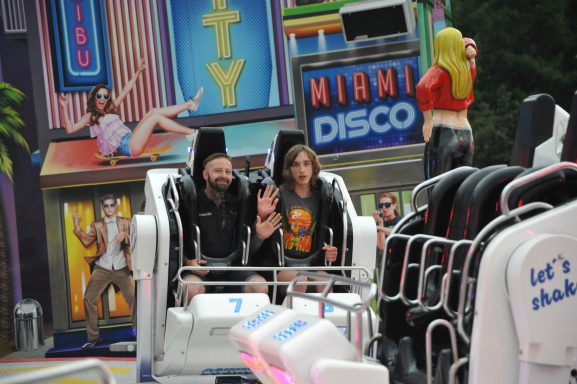Zwei Personen sitzen auf einer Achterbahn vor einem bunten Achterbahn-Backdrop.