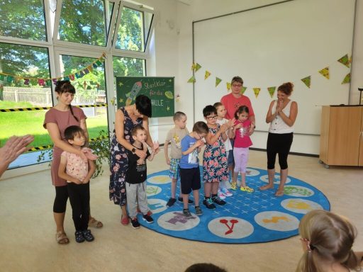 Kinder stehen auf einem bunten Teppich, umringt von Erwachsenen in einem hellen Raum.