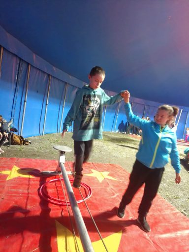 Zwei Kinder balancieren auf einem Seil im Zelt, eines hält die Hand des anderen.