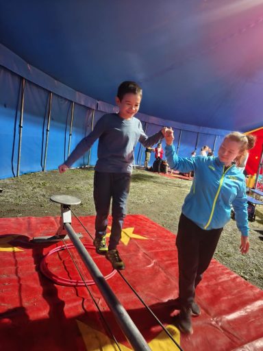 Kinder balancieren auf einem Drahtseil in einem Zirkuszelt.