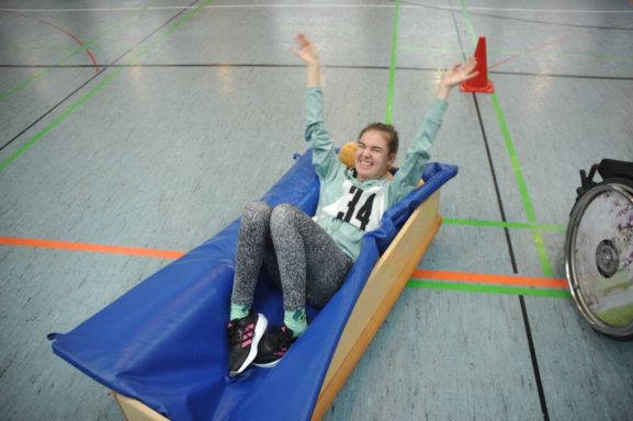 Junge Frau liegt freudig auf einer schaukelnden Spielmatte in einer Sporthalle.