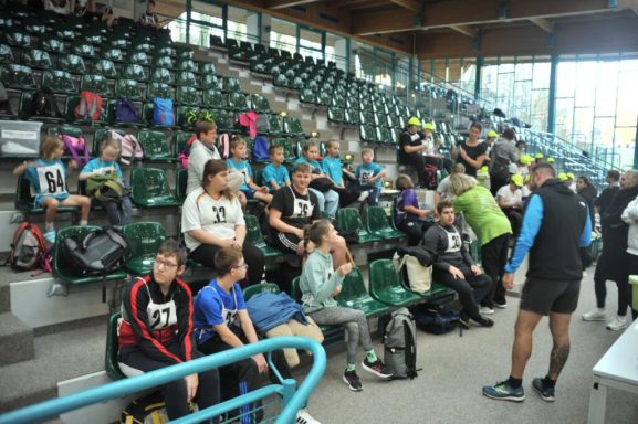 Kinder und Jugendliche sitzen in einer Sportarena auf grünen Tribünen.