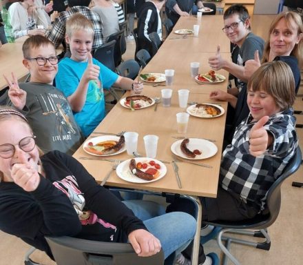 Sechs Kinder und Jugendliche sitzen an einem Tisch, zeigen Daumen hoch und genießen Essen.