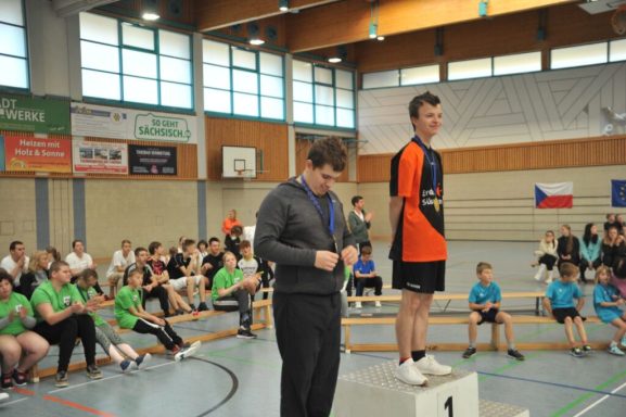 Zwei Sportler auf dem Siegerpodest, Zuschauer sitzen aufmerksam im Hintergrund.