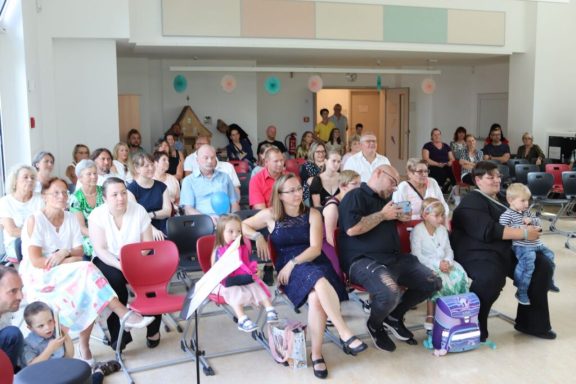 Eine Gruppe von Menschen sitzt in einem Raum, einige mit Kindern, auf Stühlen und Bänken.