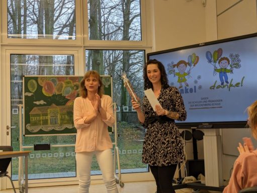 Zwei Frauen stehen im Vordergrund, eine hält ein Blatt Papier, im Hintergrund eine Präsentation.