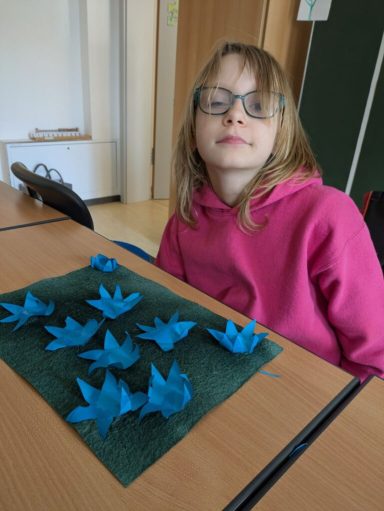 Ein Mädchen mit Brille sitzt an einem Tisch, umgeben von blauen Origami-Kranichen.