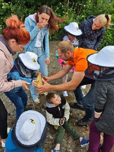 Gruppe von Menschen, die Kindern beim Umgang mit Bienen zeigen. Einige tragen Schutzhelme.