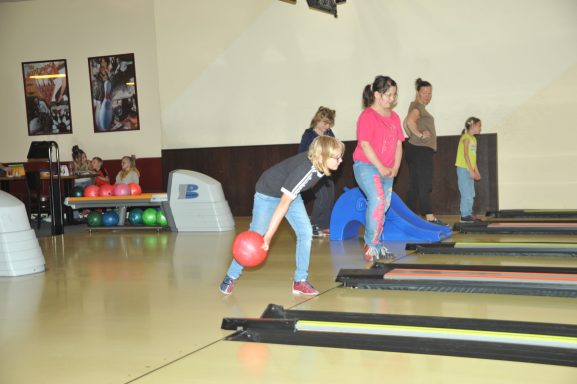 Kinder spielen Bowling, eine mit einem roten Ball hebt sich zum Wurf.