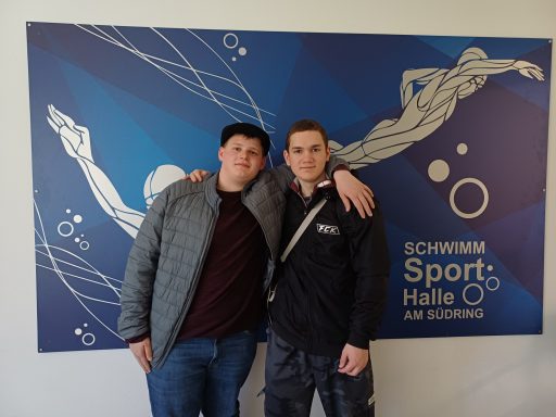 Zwei junge Männer posieren vor einem Wandbild mit Schwimmermotiv in der Schwimmhalle.