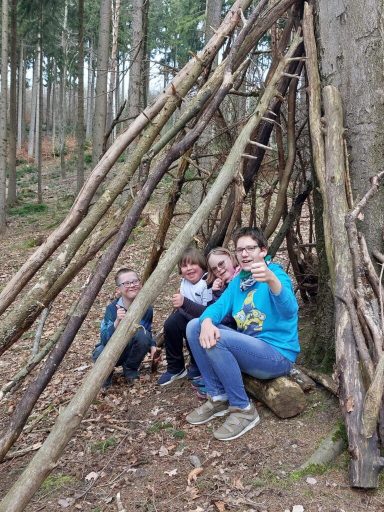 Vier Personen sitzen vor einem aus Ästen gebauten Unterschlupf im Wald.