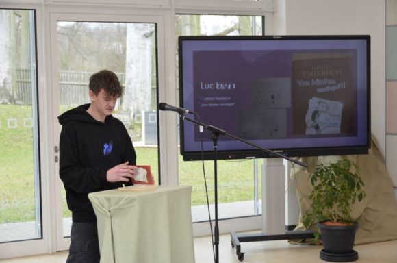 Junger Mann steht an einem Tisch mit Mikrofon vor einem Bildschirm mit Präsentation.