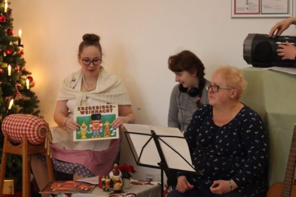 Drei Personen in festlicher Umgebung, eine hält ein buntes Buch, Weihnachtsbaum im Hintergrund.