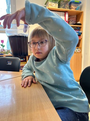 Ein Kind mit Brille hebt ein Glas mit dunklem Getränk auf einem Tisch.