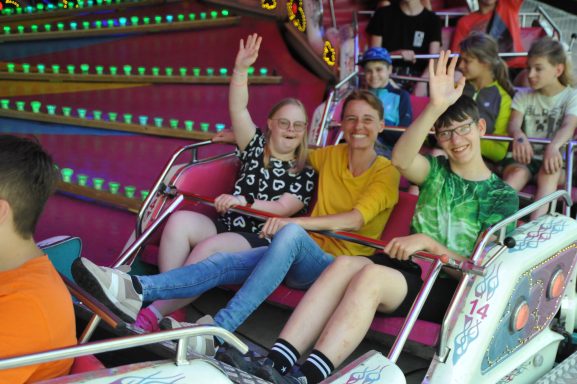 Drei lachende Jugendliche in einer Fahrgeschäfte, die fröhlich in die Kamera winken.