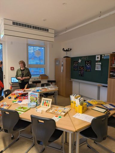 Ein Klassenzimmer mit Tischen, Lehrmaterialien und einer Tafel. Schüler und Lehrkraft anwesend.
