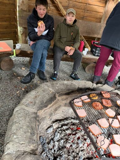 Zwei Kinder sitzen am Feuerplatz und halten Essen in der Hand. Gegrillte Scheiben sind sichtbar.