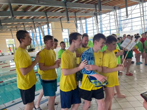 Gruppierung von Männern in gelben Shirts am Beckenrand eines Schwimmbads, fröhlich zusammen.