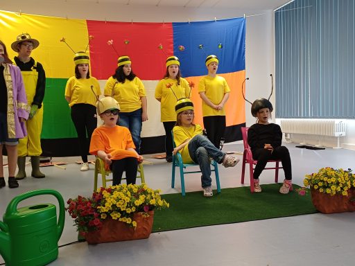 Eine Gruppe von Menschen in bunten Kostümen steht und sitzt auf einer Bühne mit Blumen.